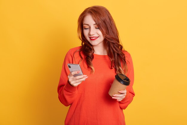 Adorabile ragazza che indossa un maglione arancione in posa isolata, tenendo il caffè da portare via e telefono cellulare