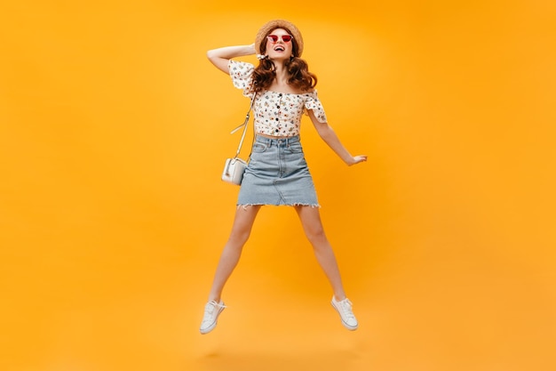 Adorabile ragazza che salta felicemente su sfondo arancione Ritratto a figura intera di giovane donna con cappello di paglia occhiali da sole rossi minigonna e camicetta con stampa floreale