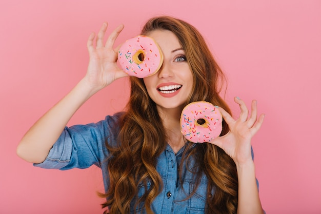 Adorabile ragazza dai capelli lunghi in camicia di jeans gioca con ciambelle glassate prima di bere il tè con gli amici. Eccitato affascinante giovane donna con i capelli ricci felice di acquistare le sue ciambelle dolci preferite in pasticceria.