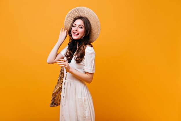 Adorabile ragazza dai capelli scuri in abito di cotone bianco sorride, tiene un cappello a tesa larga e un sacchetto di corda.