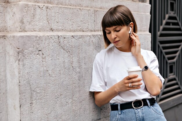 Adorabile ragazza europea con i capelli corti che indossa maglietta e jeans che ascolta musica in cuffie wireless mentre beve caffè all'aperto vicino al muro beige della città