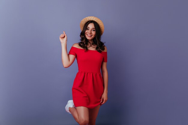 Adorabile ragazza formosa in abiti estivi alla moda ballando. Foto della signora dai capelli scuri in abito rosso in piedi nel muro viola.