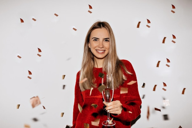 Adorabile signora affascinante con capelli chiari e trucco nudo sta sorridendo in maglione rosso con un sorriso felice su sfondo isolato sotto i coriandoli