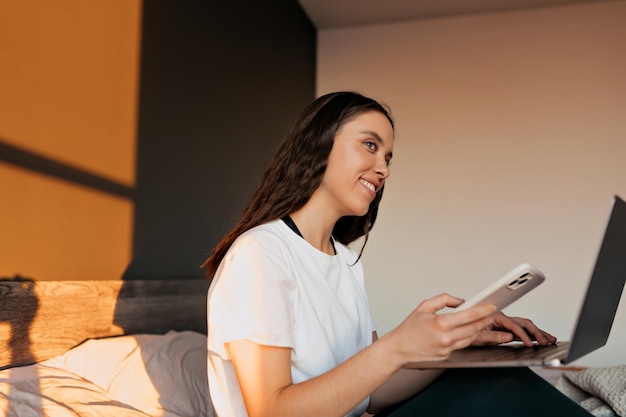 Adorabile signora carina in abiti da casa sta scorrendo lo smartphone e guardando la finestra alla luce del sole del mattino La donna sta lavorando con il laptop da casa nel letto