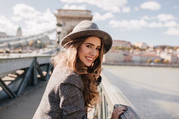 Adorabile signora con lunghi capelli castano chiaro in posa su brige su sfocatura dello sfondo della città