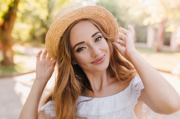 Adorabile signora con occhi verdi felici che sorridono dolcemente godendo del clima caldo nel parco. Foto all'aperto del primo piano della ragazza felice che tiene il cappello di paglia con entrambe le mani di buon umore, camminando sulla strada.