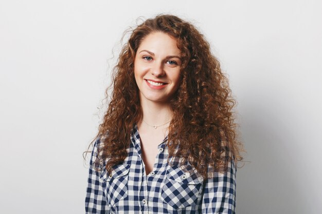 Adorabile studentessa piuttosto giovane con i capelli lunghi ricci e il sorriso caldo guarda con gioia la fotocamera