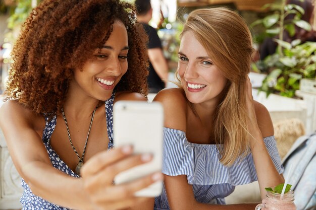 Adorabili donne di diverse nazionalità posano per selfie e si divertono insieme, hanno espressioni positive, usano le moderne tecnologie per intrattenersi, postano foto da scaricare in rete