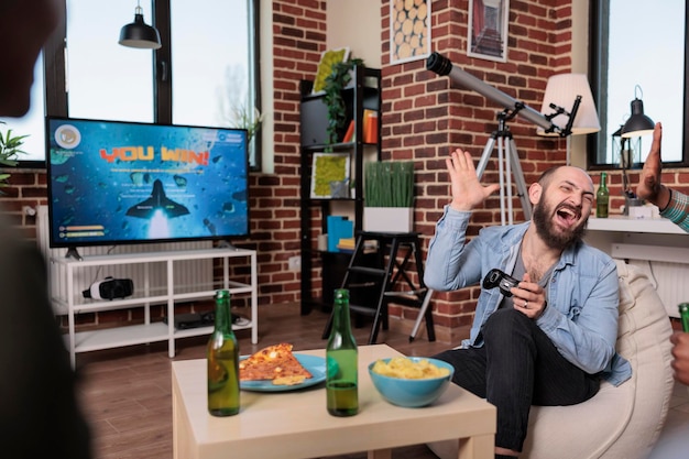 Adulti felici che celebrano i videogiochi vincono sulla console TV, giocando a gare di tiro con un gruppo di amici al raduno. Divertirsi e vincere al gioco in televisione, attività ricreative.