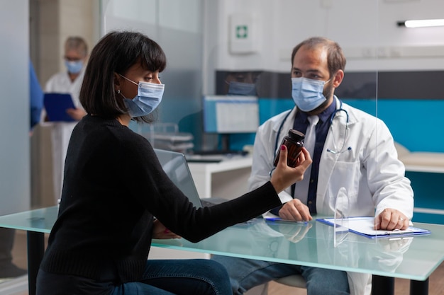 Adulto e medico con maschera facciale che discutono della bottiglia di pillole e del trattamento di prescrizione durante la visita di controllo. Medico generico che somministra medicinali al paziente per curare la malattia durante la pandemia.