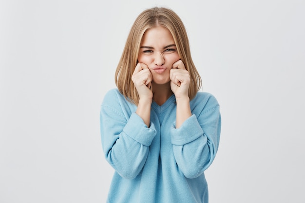 Affascinante, accigliata sul viso, giovane donna europea con i capelli biondi che indossa un maglione blu, pizzicandole le guance, beffardo, di buon umore e divertimento.