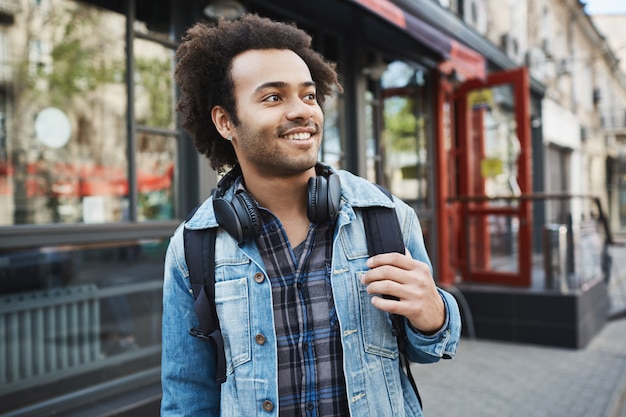 Affascinante bel maschio afro-americano con setola e taglio di capelli afro che osserva da parte mentre si tiene lo zaino e passeggiando in città.