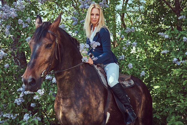 Affascinante bella bionda fantino che monta un cavallo marrone nel giardino fiorito.