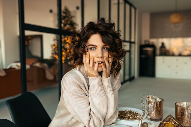 Affascinante bella donna con acconciatura ondulata camicia in posa con emozioni sorprese mentre era seduto alla tavola di Natale sopra l'albero di Natale