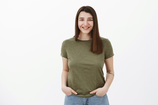 Affascinante bella donna europea con capelli castani e tatuaggio sorridente tenera e intrattenuta mentre posa sul muro bianco, tenendo le mani in tasca sentendosi gioiosa e ottimista mentre parla piacevolmente