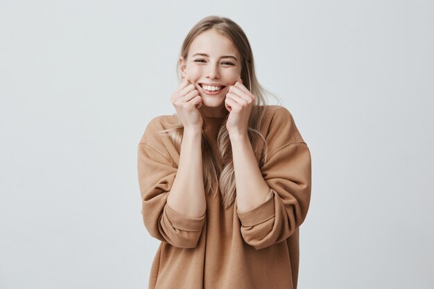 Affascinante bellissimo modello femminile che sorride ampiamente indossando un maglione marrone, pizzicandole le guance, beffardo, avendo buon umore e divertimento. Emozioni e sentimenti positivi