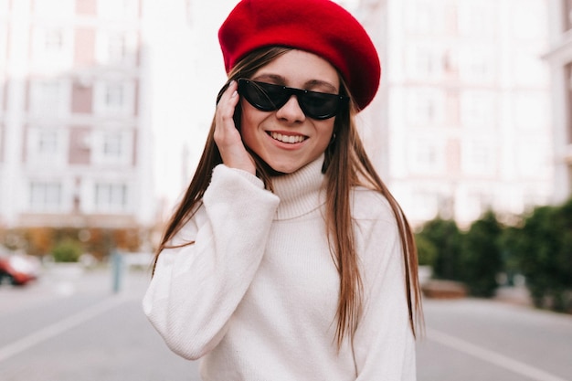 Affascinante donna abbronzata bruna con berretto rosso da vista e pullover bianco sorride ampiamente e posa fuori in un caffè di strada