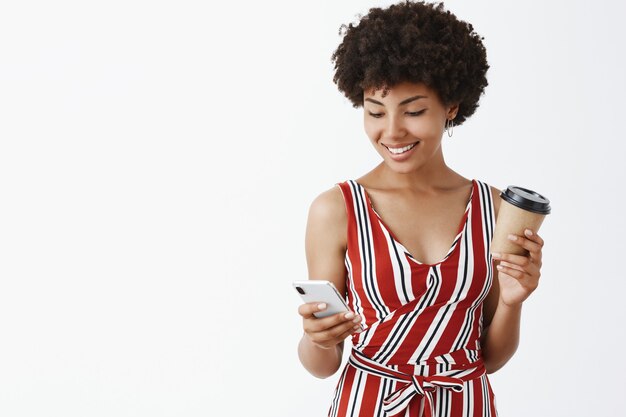 Affascinante donna afroamericana moderna femminile in vestito elegante che tiene tazza di carta e smartphone che sorride allo schermo che gode del buongiorno