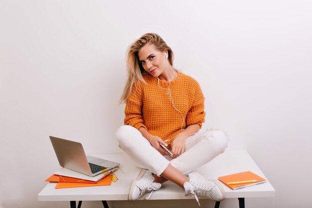 Affascinante donna bionda in scarpe sportive seduto sulla scrivania e guardando con un sorriso giocoso dopo il lavoro con il computer portatile