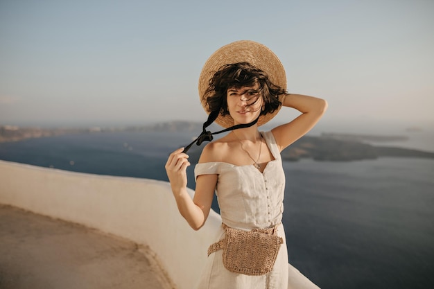 Affascinante donna bruna riccia tiene cappello di paglia e sorride Bella signora in abito beige elegante posa sul posto con vista mare