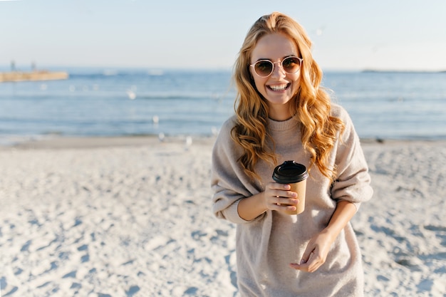 Affascinante donna con capelli ondulati, bere il tè in spiaggia. Donna alla moda in maglione rilassante in una giornata autunnale in spiaggia.