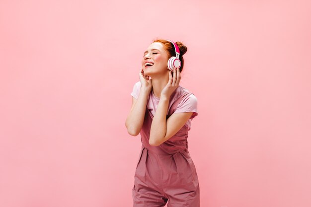Affascinante donna con un sorriso esamina la macchina fotografica, tenendo lo smartphone nelle sue mani. donna in abito rosa ascoltando musica in cuffia.