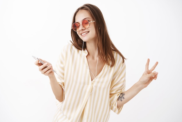 Affascinante donna europea gentile in occhiali da sole e camicetta gialla elegante ascoltando musica in auricolari che tengono smartphone ballando mostrando gesto di pace godendo del suono
