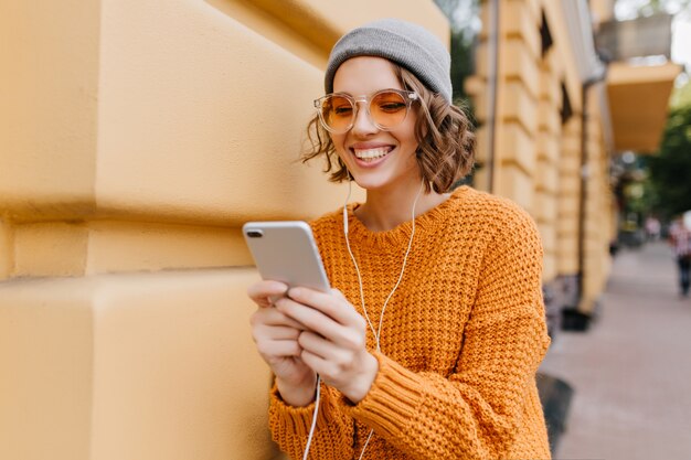 Affascinante donna femminile in cappello e auricolari sorridente durante la videochiamata con un amico