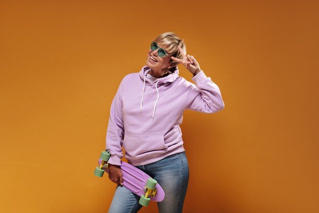 Affascinante donna fresca con l'acconciatura moderna bionda in occhiali verdi e felpa con cappuccio oversize rosa sorridente, che mostra il segno di pace e in posa con lo skateboard su sfondo arancione.