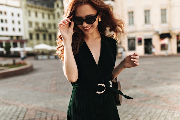 Affascinante donna in abito di velluto e occhiali da sole sorridente all'esterno