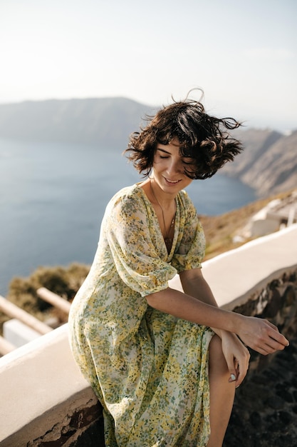 Affascinante donna in abito floreale sorride sinceramente fuori Bruna riccia signora in elegante abito estivo si siede sullo sfondo di montagna e mare