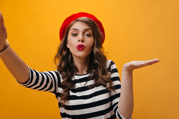 Affascinante donna in camicia a righe e berretto soffia bacio e prende selfie. Bella ragazza con cappello rosso brillante in camicetta moderna che esamina la macchina fotografica.