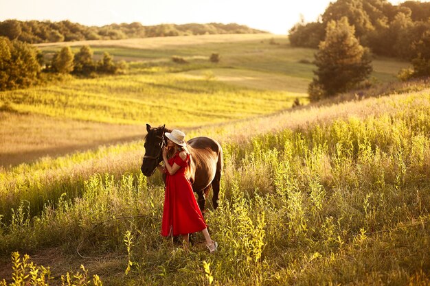 Affascinante donna in un cappello di fieno e vestito rosso si leva in piedi con un cavallo sul campo verde