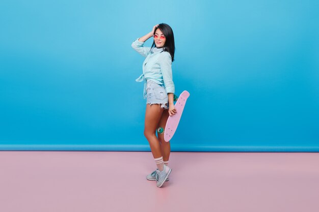 Affascinante donna ispanica con capelli neri dritti in piedi sulla punta dei piedi e tenendo lo skateboard rosa. La foto dell'interno del modello femminile abbastanza asiatico in occhiali da sole indossa scarpe sportive e pantaloncini di jeans.