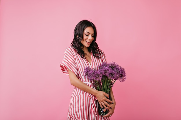 Affascinante donna latina con affetto guarda il bouquet di fiori selvatici viola. Ragazza in vestito a strisce in posa imbarazzata.