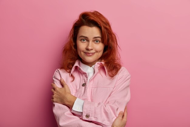 Affascinante donna naturale si abbraccia, ha un'alta autostima, indossa una giacca alla moda in denim rosa, fossette sulle guance, capelli voluminosi