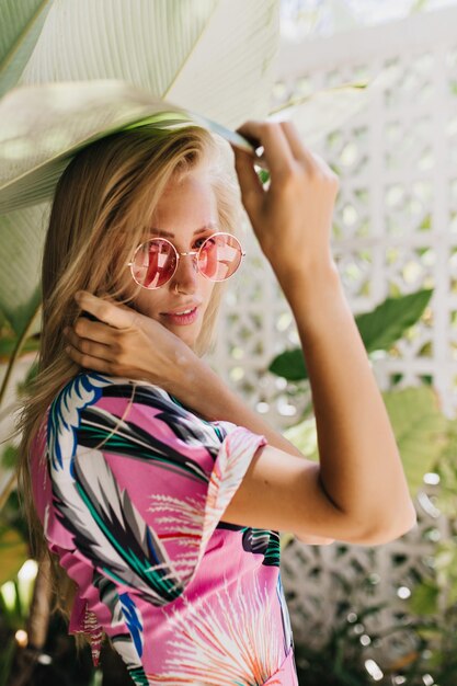 Affascinante donna snella con la pelle abbronzata che guarda sopra la spalla durante il servizio fotografico Foto di allegra ragazza bionda con occhiali da sole rosa in piedi vicino a piante verdi