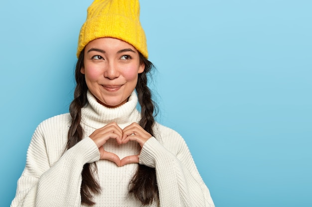 Affascinante e felice giovane donna asiatica fa il segno del cuore, confessa il fidanzato innamorato, ha l'espressione del viso compiaciuto, guarda da parte, indossa un cappello e un maglione gialli