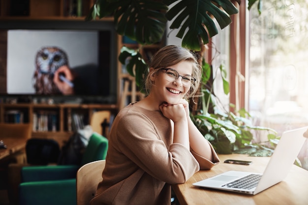 Affascinante e spensierata donna europea con capelli biondi in occhiali, seduta vicino alla finestra e laptop, appoggiata sulle mani