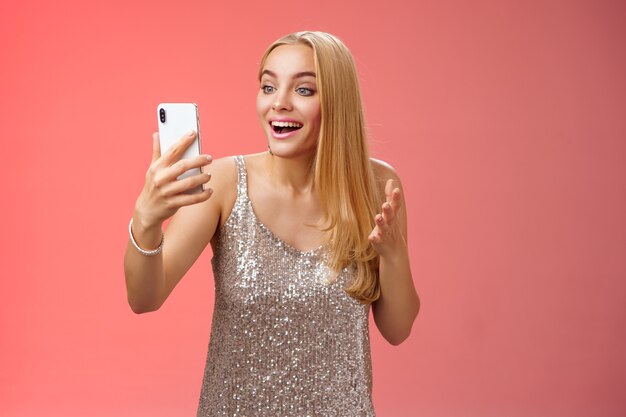 Affascinante elegante bella ragazza bionda in abito d'argento parlando videochiamata parlando guardando il display dello smartphone divertito sorpreso sorridente felicemente avere conversazione fratello che mostra vestito da ballo