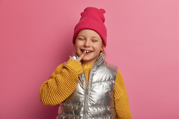 Affascinante figlia piccola, piccola bambina indica il suo nuovo dente, sorride ampiamente, indossa cappello, maglione e gilet lavorato a maglia, previene la carie, si prende cura dei denti, modella su un muro rosa pastello