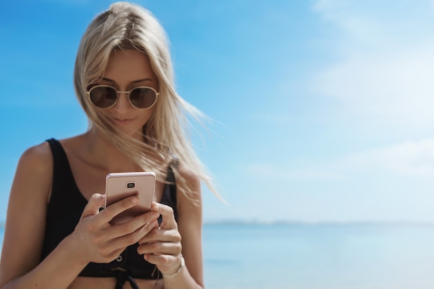 Affascinante giovane donna che gode delle vacanze estive nella soleggiata spiaggia sabbiosa utilizzando il cellulare