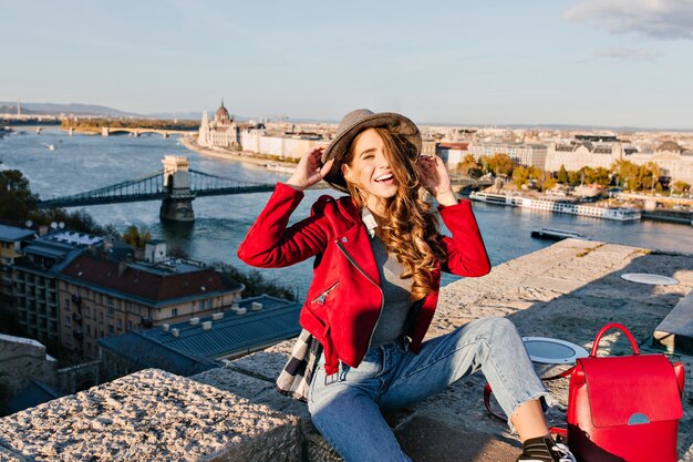 Affascinante giovane donna con capelli castani tenendo il cappello e ridendo sullo sfondo della città