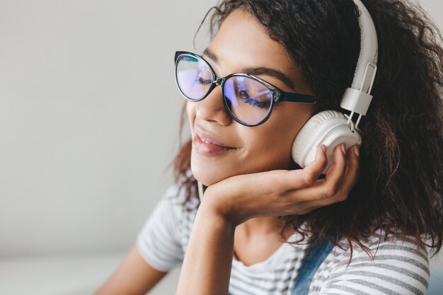 Affascinante giovane donna con la pelle marrone e lunghe ciglia nere che si gode la musica preferita in grandi auricolari