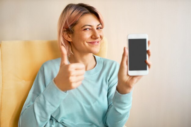 Affascinante giovane donna felice con capelli rosati che tiene smart phone con display nero vuoto con copia spazio per il contenuto pubblicitario, facendo il pollice in alto gesto come segno di appoval, ammiccando alla telecamera