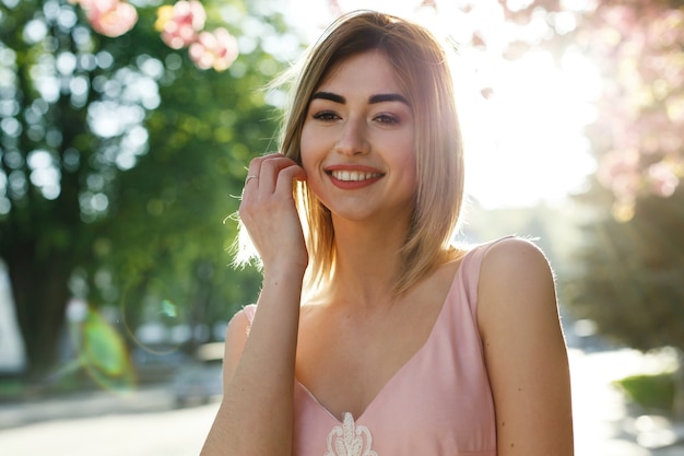 Affascinante giovane donna in abito rosa pone davanti a un albero di sakura pieno di fiori rosa