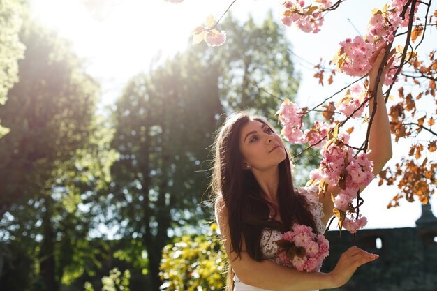 Affascinante giovane donna in abito rosa pone davanti a un albero di sakura pieno di fiori rosa