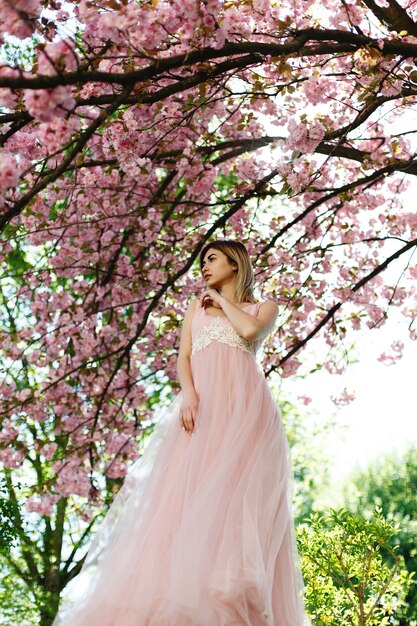 Affascinante giovane donna in abito rosa pone davanti a un albero di sakura pieno di fiori rosa