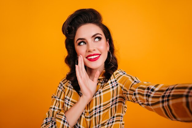 Affascinante giovane donna in camicia a scacchi prendendo selfie. Studio shot di sorprendente ragazza pinup isolata su sfondo giallo.