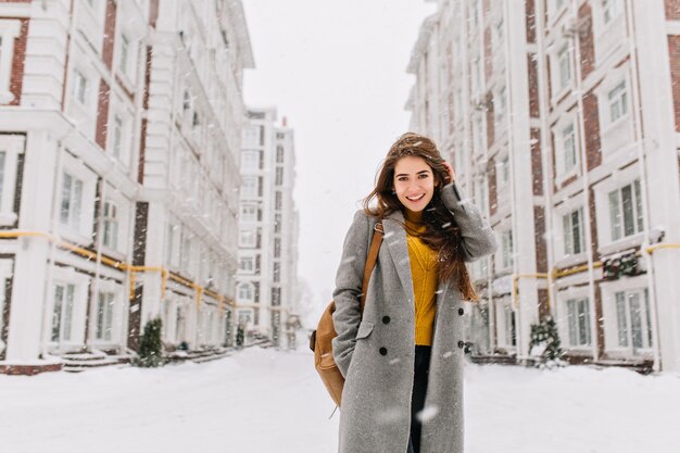 Affascinante giovane donna in cappotto con capelli lunghi del brunette che gode della nevicata in una grande città. Emozioni allegre, sorridente, atmosfera natalizia, emozioni positive del viso, clima invernale. Posto per il testo.